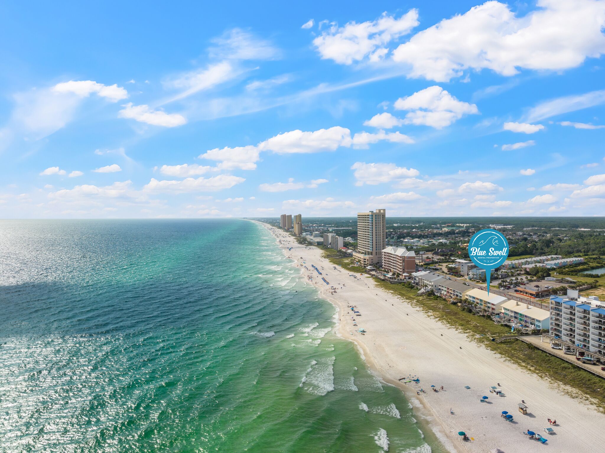 Un paysage urbain côtier avec une plage, des hôtels et un panneau indiquant « Blue Swell Rental Properties ».