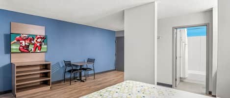 Habitación de hotel con cama, televisor montado en la pared, escritorio con sillas y zona de ducha.