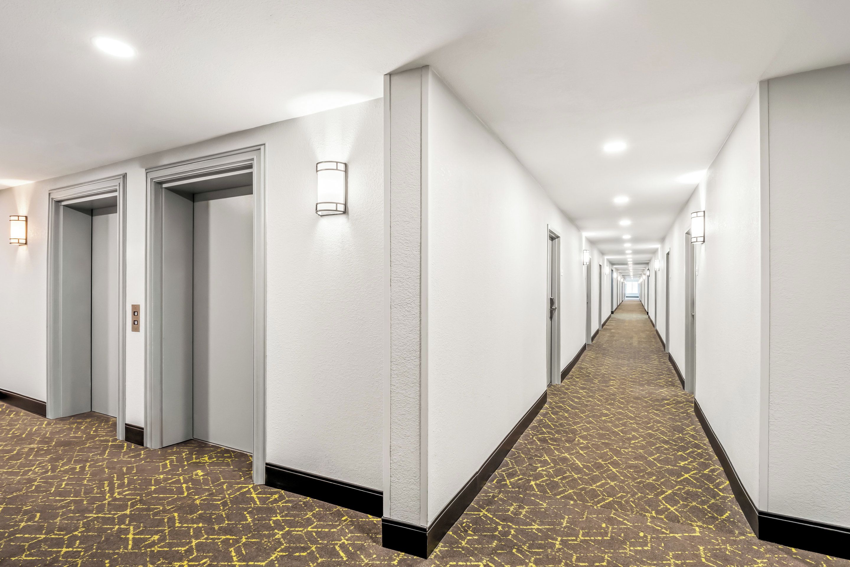 A long, narrow hotel corridor with white walls, recessed lighting, and patterned carpet.