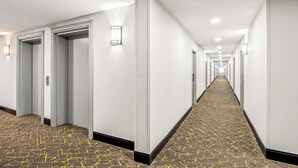 A long, narrow hotel corridor with white walls, recessed lighting, and patterned carpet. - HomeTowne Studios by Red Roof Southfield (Southfield)