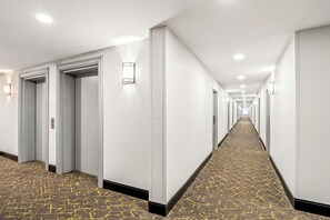 A long, narrow hotel corridor with white walls, recessed lighting, and patterned carpet. - HomeTowne Studios by Red Roof Southfield (Southfield)