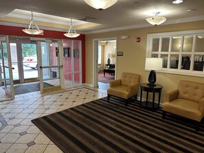 A hotel lobby with a check-in desk, two armchairs, a patterned rug, and a large window. - HomeTowne Studios by Red Roof Southfield (Southfield)