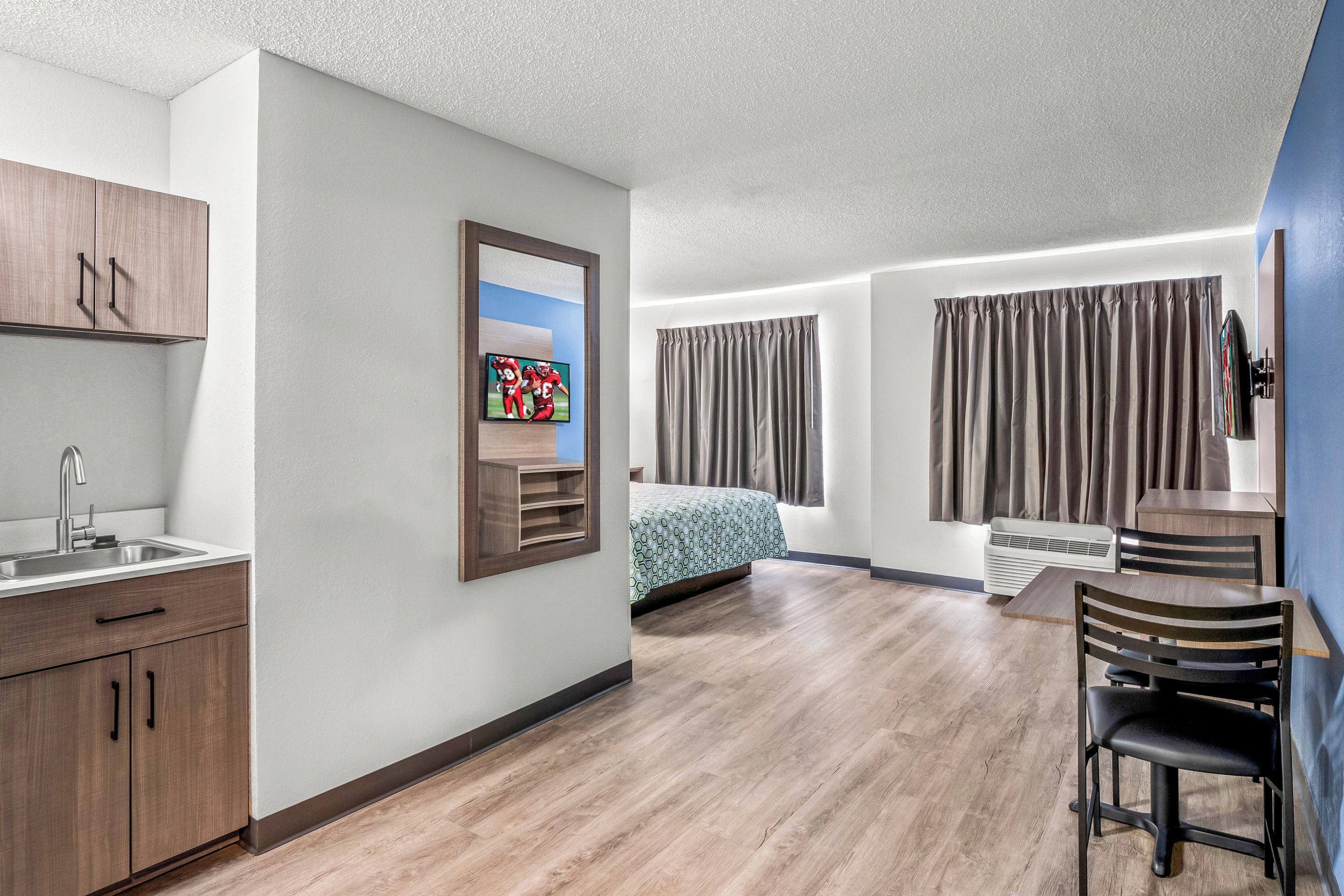 Een moderne hotelkamer met een houten vloer, een bed met een decoratief sprei, een bureau met stoel, een aan de muur bevestigde televisie en een raam met gordijnen.