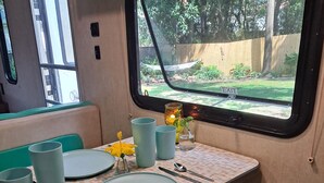 Una kitchenette compacta con vista al jardín por la ventana, una mesa puesta con platos, tazas y utensilios, y un banco con cojín.