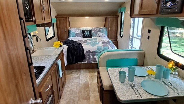 Interior compacto de autocaravana con kitchenette, una cama con ropa de cama estampada, zona de comedor con mesa para dos personas y un tragaluz sobre la cama.