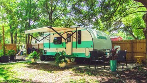 Una caravana estacionada en un patio trasero, con área de patio, rodeada de árboles y plantas en macetas.