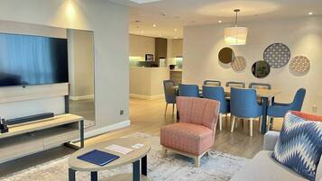 Una sala de estar moderna con un televisor de pantalla plana, una zona de comedor con sillas azules y una cocina al fondo.