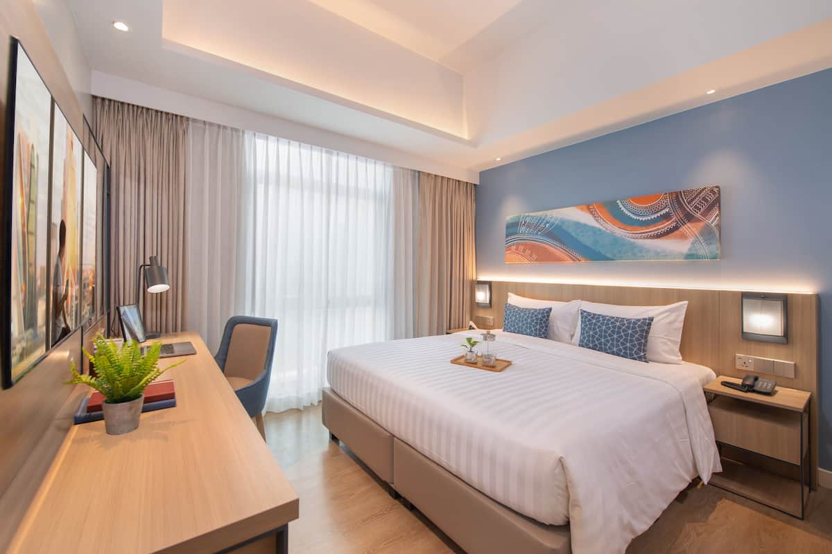 A modern hotel room with a large bed, a desk with a plant, a chair, and a painting on the wall.