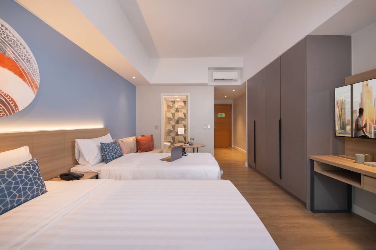 A hotel room with two beds, a desk, a TV, and a bathroom visible through a glass door.