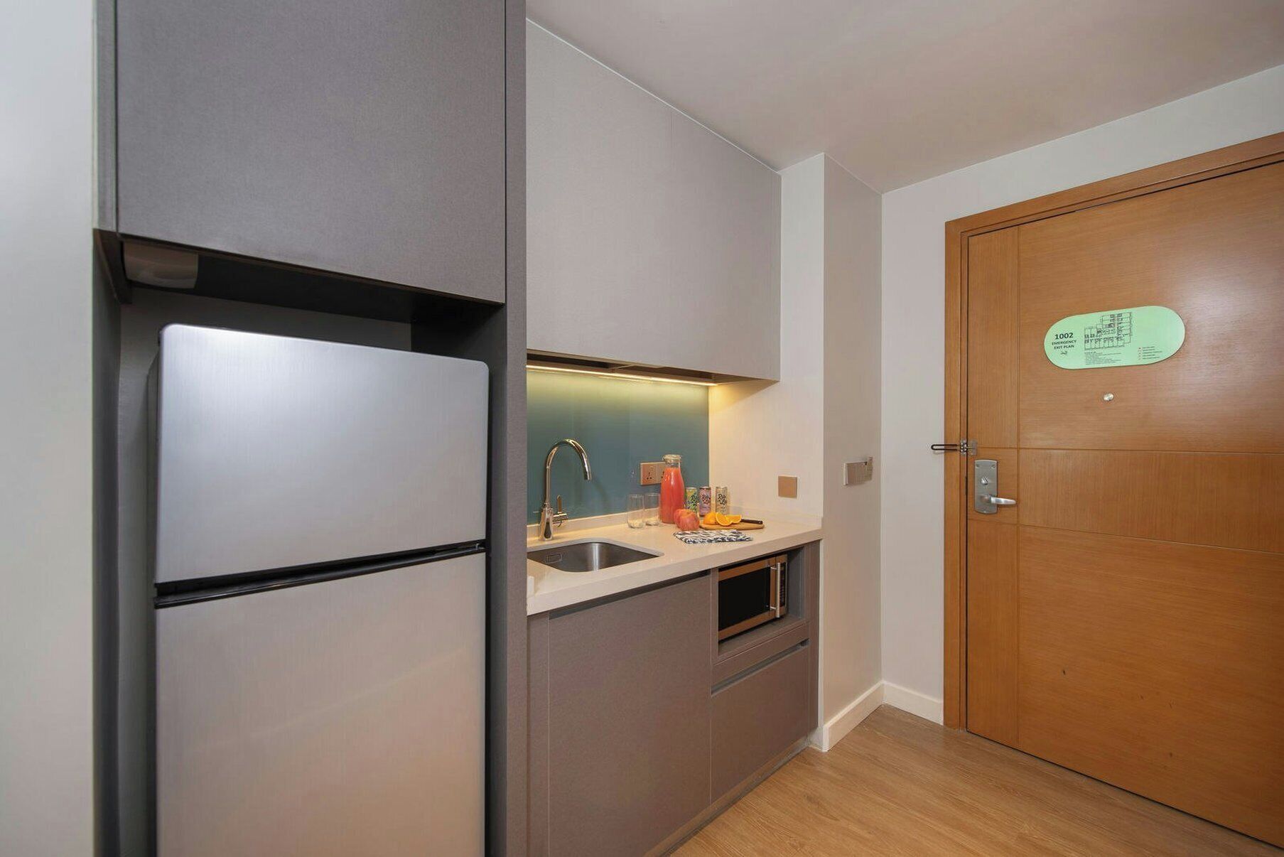Cocina compacta con refrigerador, lavaplatos y una puerta de madera con señal de salida de emergencia.