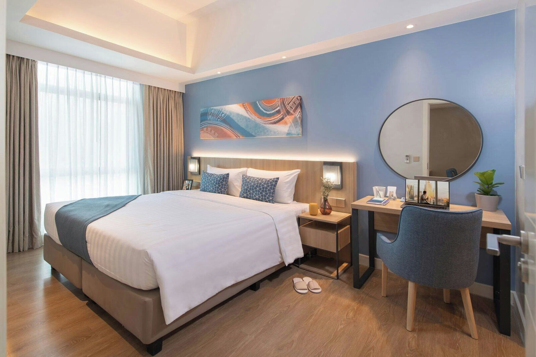 A modern hotel room with a large bed, a desk, a chair, a mirror, and a large window with curtains.