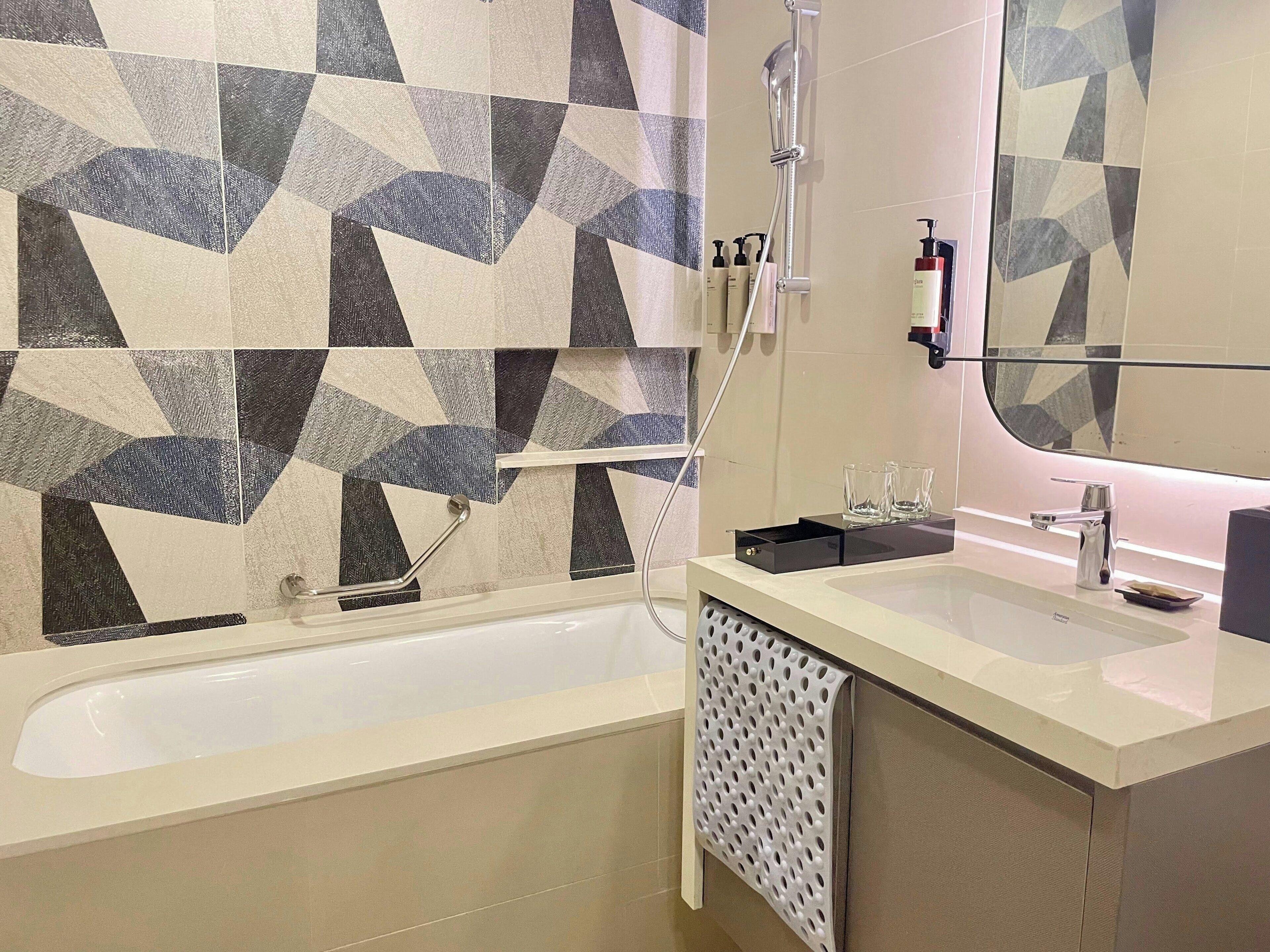 Un baño moderno con un revestimiento de azulejos geométricos, una bañera, un tocador con espejo y un perchero para toallas.