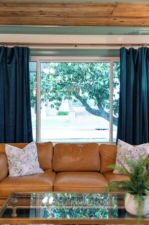 A living room with a brown leather sofa, a glass coffee table, and a view of a tree through a window with dark curtains.