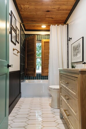 A bathroom with a bathtub, toilet, wooden vanity, and a window with shutters.