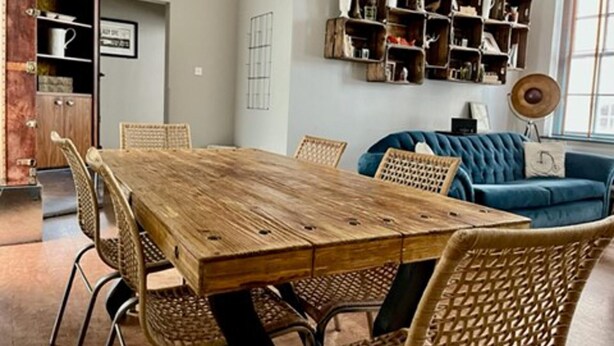 A dining area with a wooden table and wicker chairs, a blue sofa, and built-in wooden shelves with various items. - Chester Centre 2-bed Apartment -by Oddfellows (Chester)