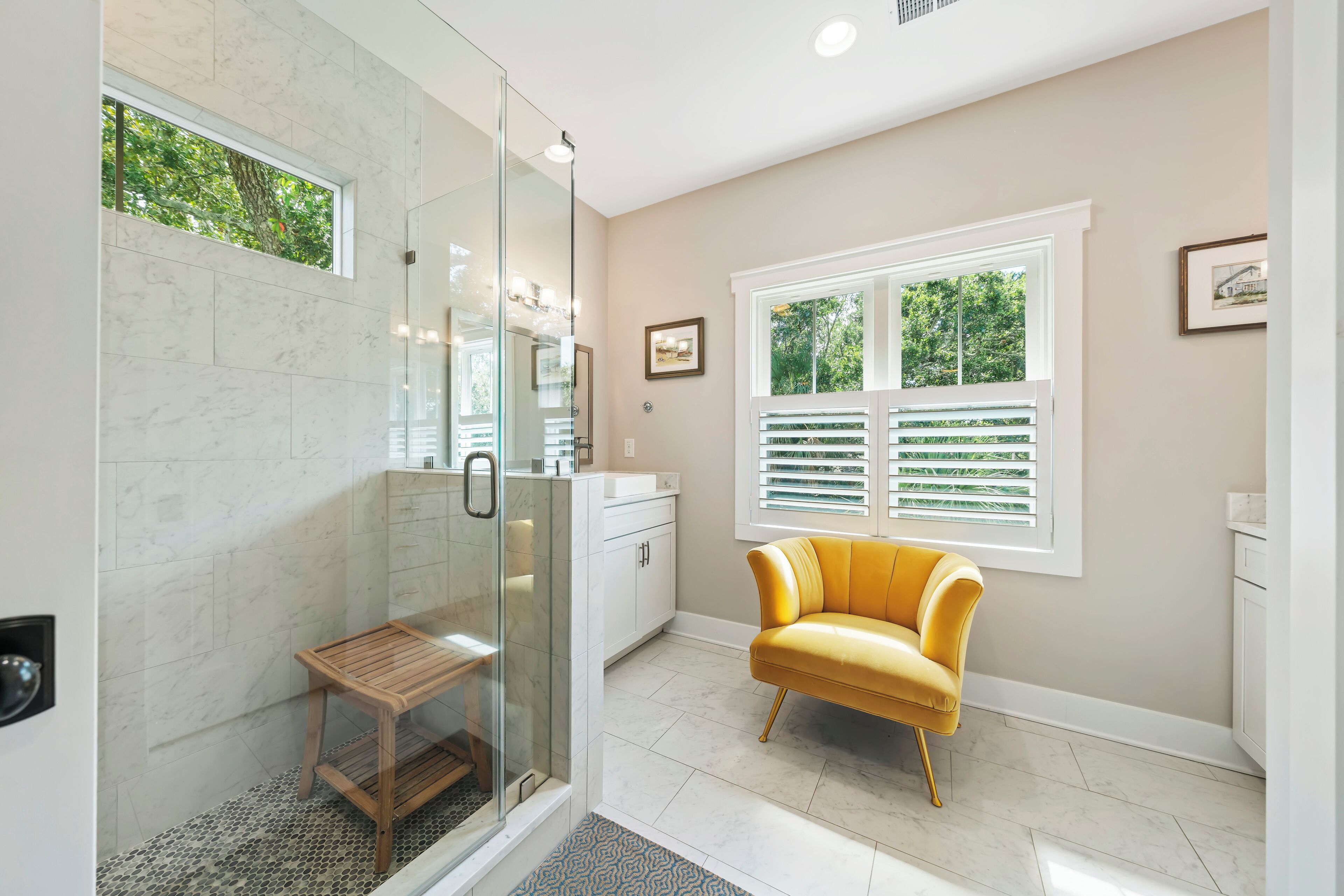 A bathroom with a glass shower enclosure, a wooden bench, a yellow armchair, and a window with blinds.