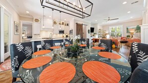 A modern dining area with a glass table surrounded by chairs with orange accents, a geometric ceiling light fixture, and a view into the adjacent kitchen and living spaces.