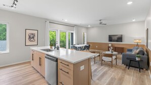 Een moderne keuken met een centraal eiland, houten kastjes en een vaatwasser. Er is een eethoek met een tafel en stoelen, en een woonkamer met een bank en een televisie.