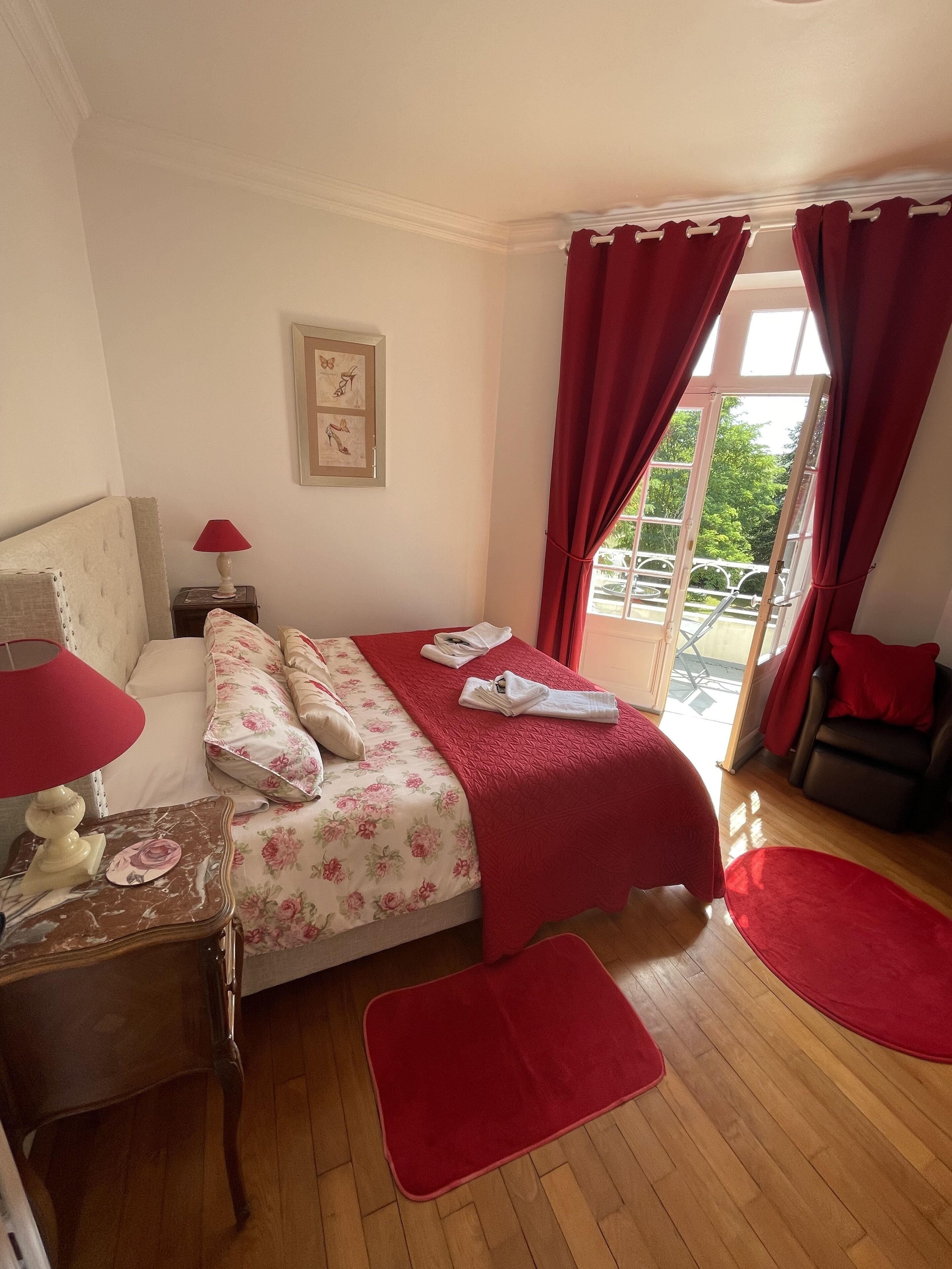 A bedroom with a bed, red curtains, a window, a wooden floor, and a small table with a lamp.