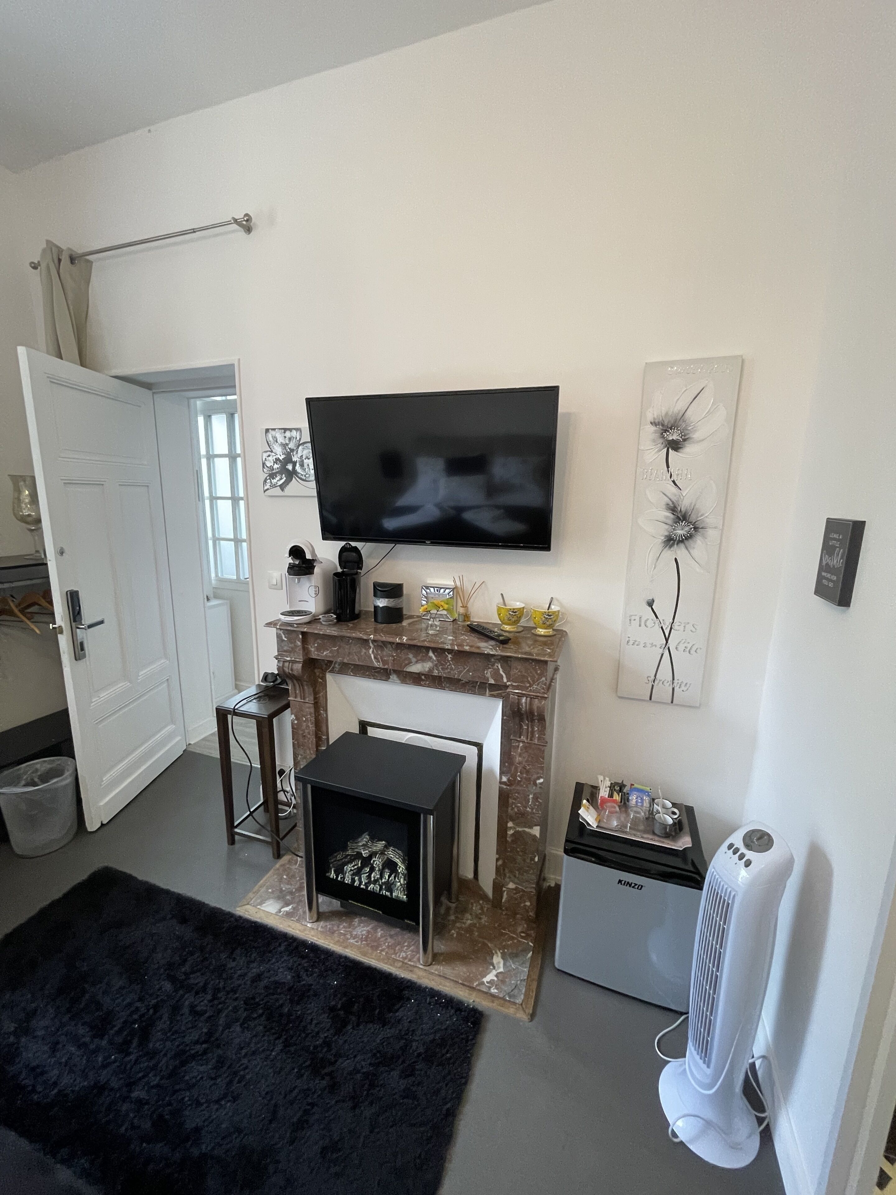 Una sala de estar con un televisor de pantalla plana sobre una chimenea, una mesita con un objeto decorativo, una alfombra negra y un ventilador blanco.
