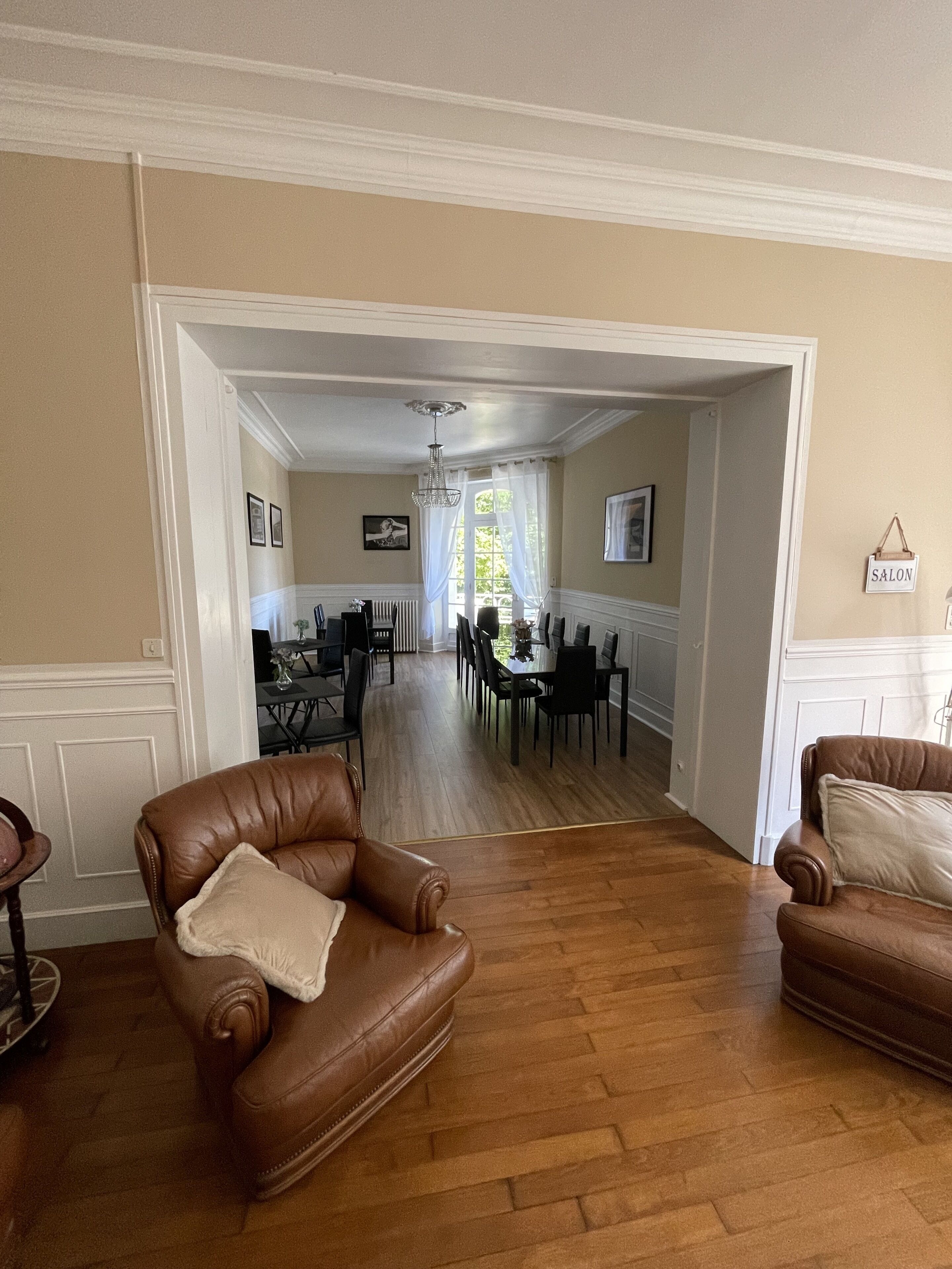 A spacious living room with a wooden floor, two leather armchairs, a dining area with a chandelier, and a 'SALON' sign on the wall.