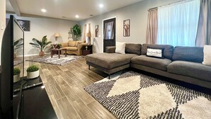 A modern living room with a sectional sofa, a coffee table, and a TV. There are two side tables with lamps and a potted plant. The room has a tiled floor and a large window with curtains. - Super Spacious Safari Retreat.. Lots of SPACE-Deck + Grill,  near Memphis/Desoto (Southaven)