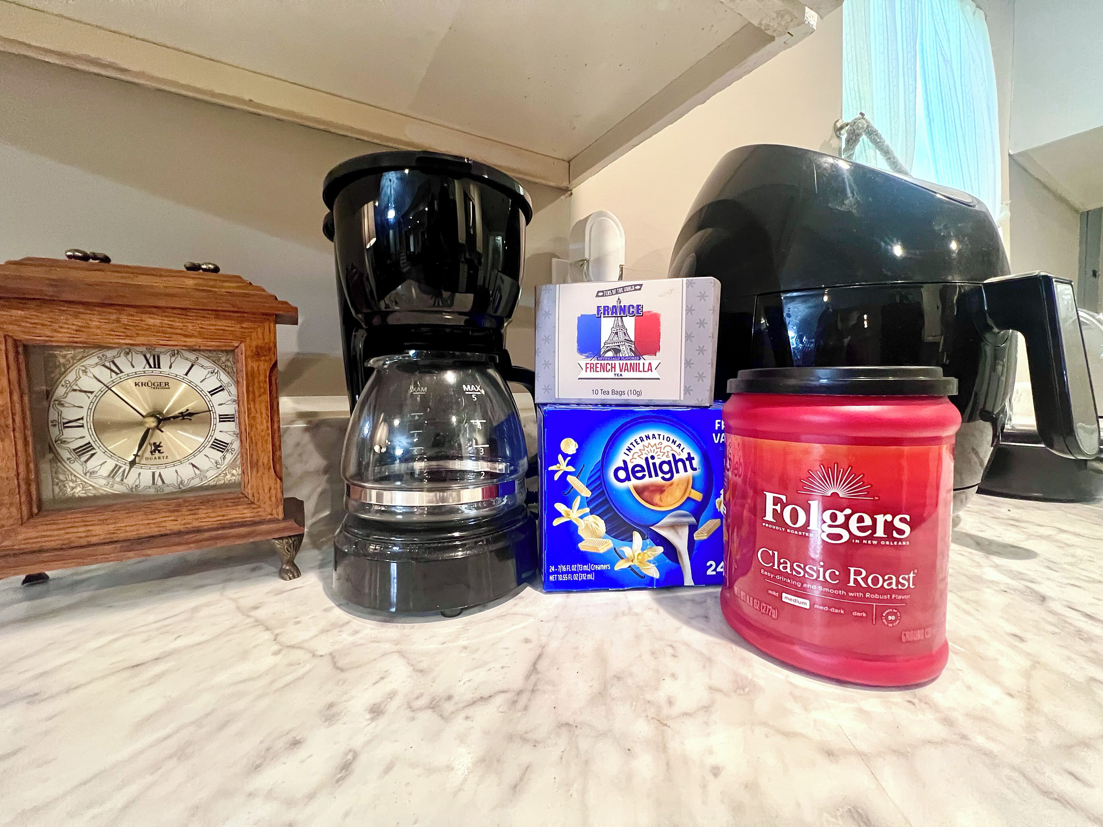 A kitchen counter with a coffee maker, a box of tea bags, a jar of Folgers coffee, and a vintage clock.