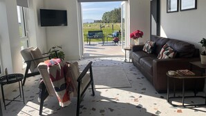 A living room with a sofa, armchair, and a television. There is a door leading outside to a patio area with a view of a green field and a bench.