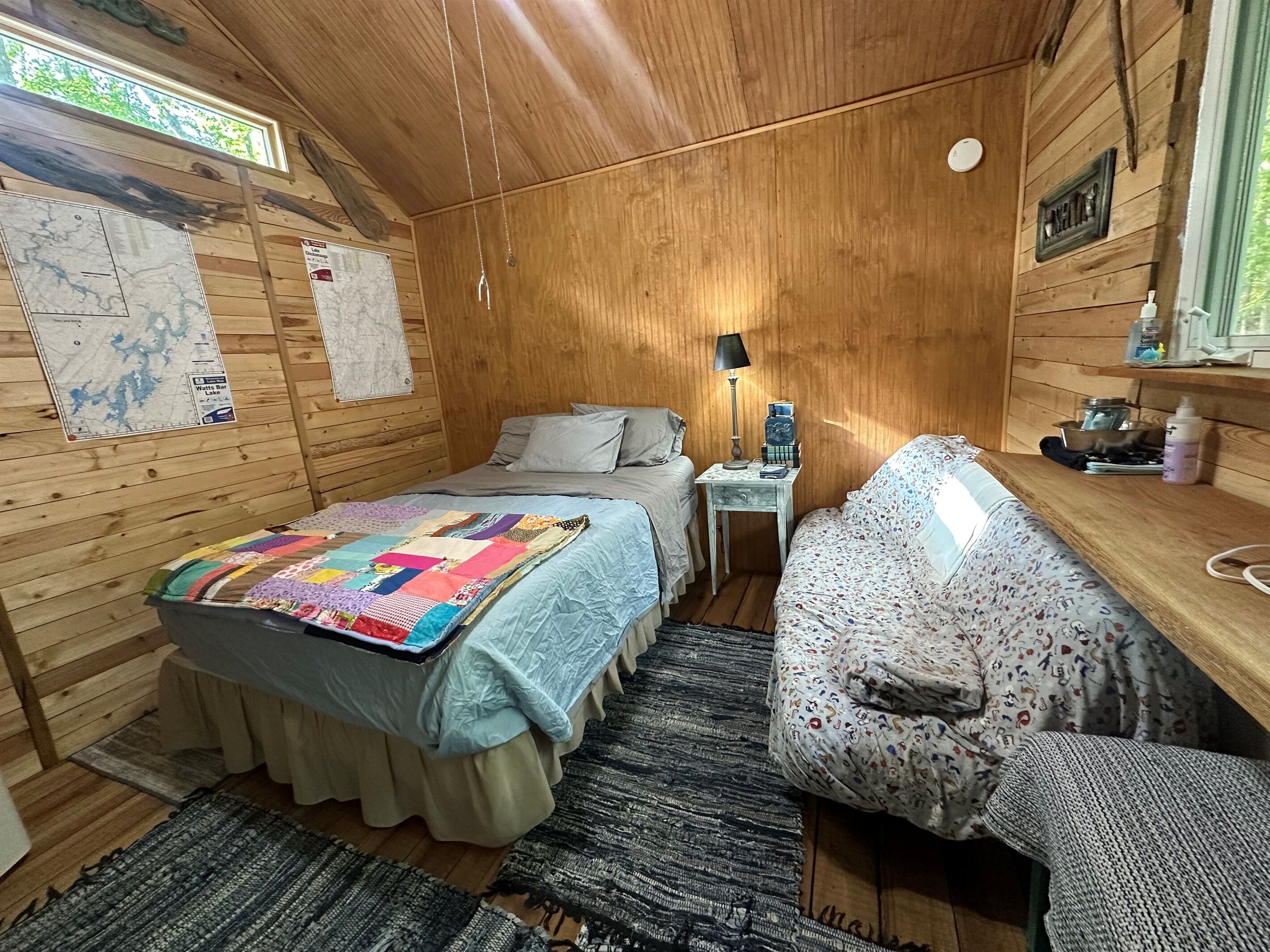 A cozy cabin room with a bed, a small table with a lamp, a map on the wall, and a window with a view.