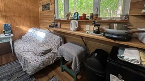 Interior de una cabaña con una cama, una pequeña mesa, una ventana con vistas a árboles y una zona de kitchenette.