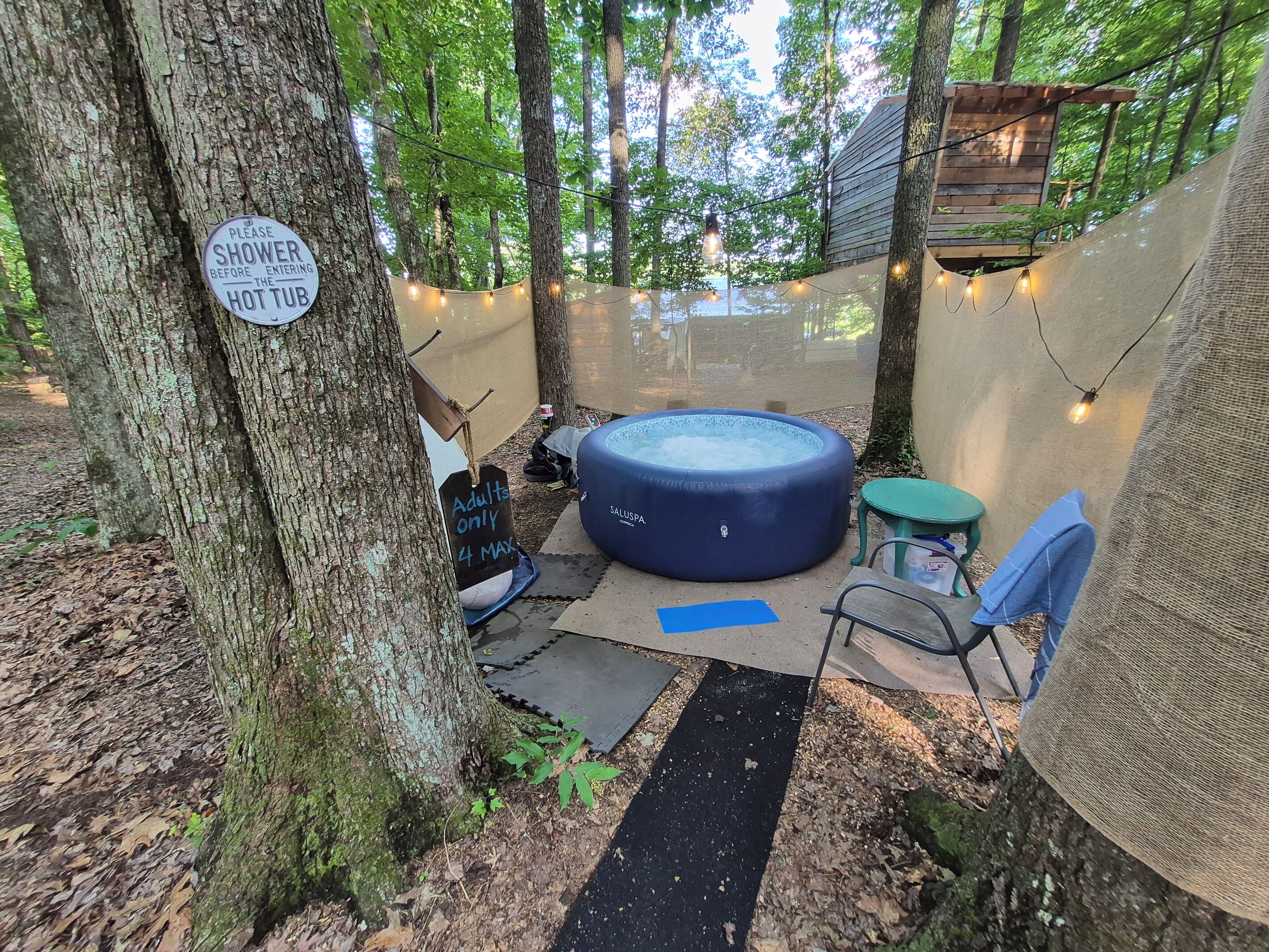 Una vasca idromassaggio circondata da alberi, un cartello che invita a fare la doccia prima di entrare e un’area relax con tavolo e sedie.
