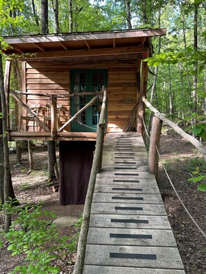 Une cabane en bois avec une terrasse en bois menant à une porte verte, le tout entouré d’arbres.