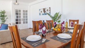 A dining area with a wooden table set for four, surrounded by wooden chairs. There is a fruit basket on the table, a bottle of wine, and a painting on the wall. - Spacious villa with BBQ and pool in Albufeira (Albufeira)