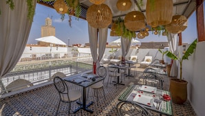 A rooftop terrace with tables and chairs, hanging wicker lamps, and a view of a tower in the background. - Riad Safran et Soleil (Marrakech)