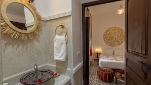 A bathroom with a round mirror, a sink with rose petals, a towel rack, and a bed with a patterned headboard. - Riad Safran et Soleil (Marrakech)