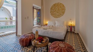A bedroom with a bed, two ottomans, a bedside table with a lamp, a decorative wall piece, and a view of a balcony with a railing. - Riad Safran et Soleil (Marrakech)