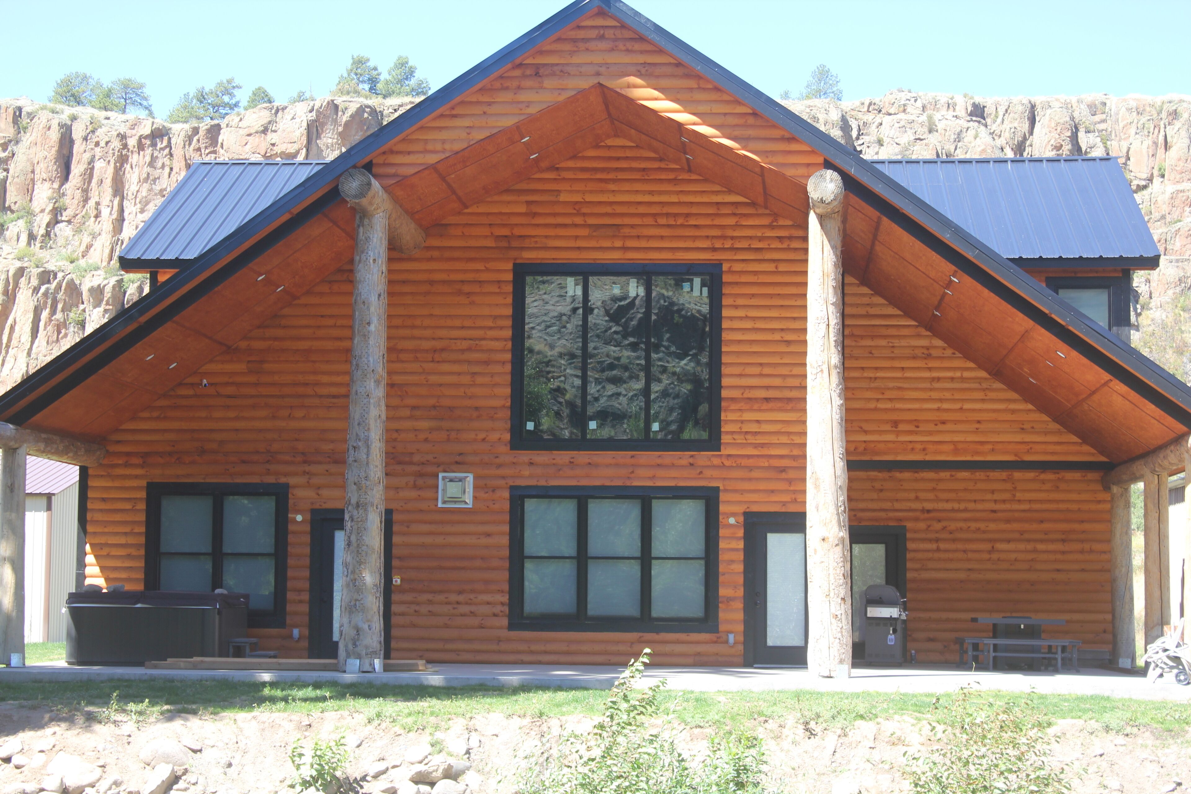 A log cabin with a large central window, a covered patio area, and a mountain backdrop.