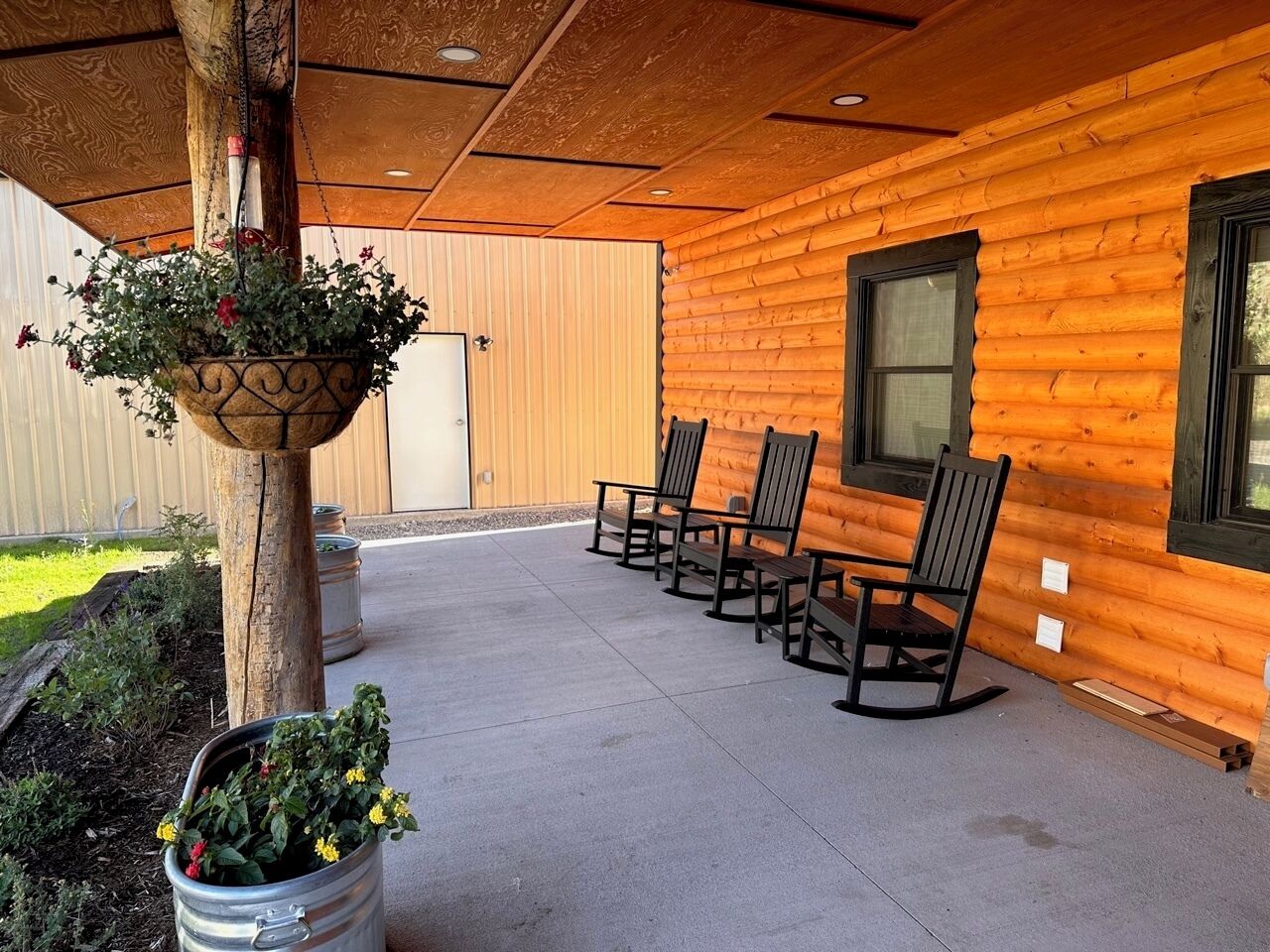 Una baita di legno con patio in cemento, quattro poltrone a dondolo e cesti di fiori pensili.