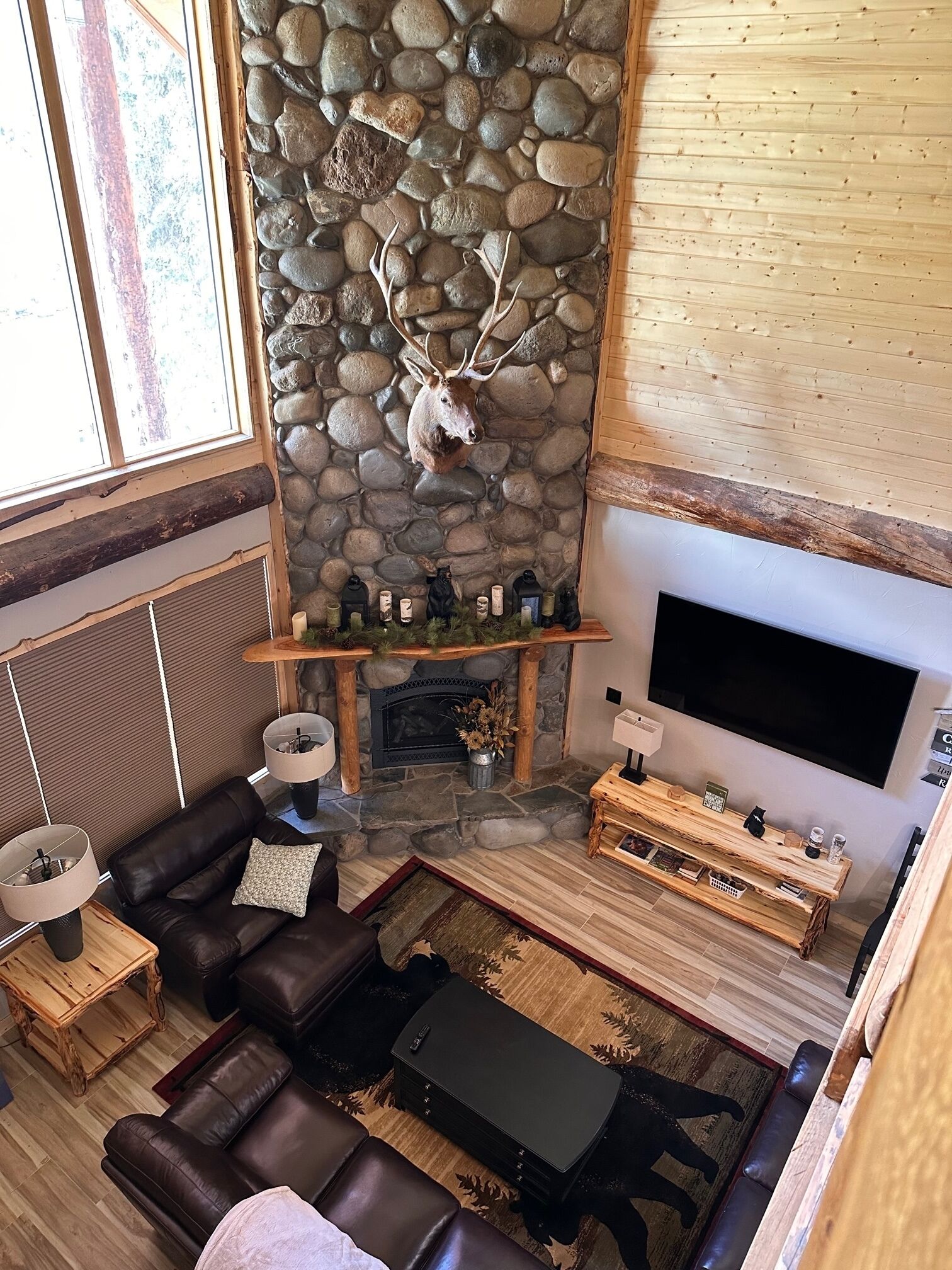 A cozy living room with a stone fireplace, leather sofas, a flat-screen TV, and a stone wall with a mounted deer head.