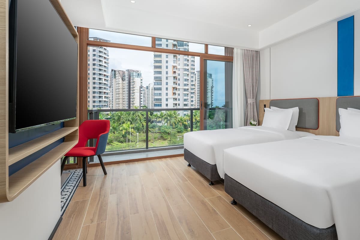A hotel room with two beds, a TV, a red chair, and a large window with a view of high-rise buildings and greenery.