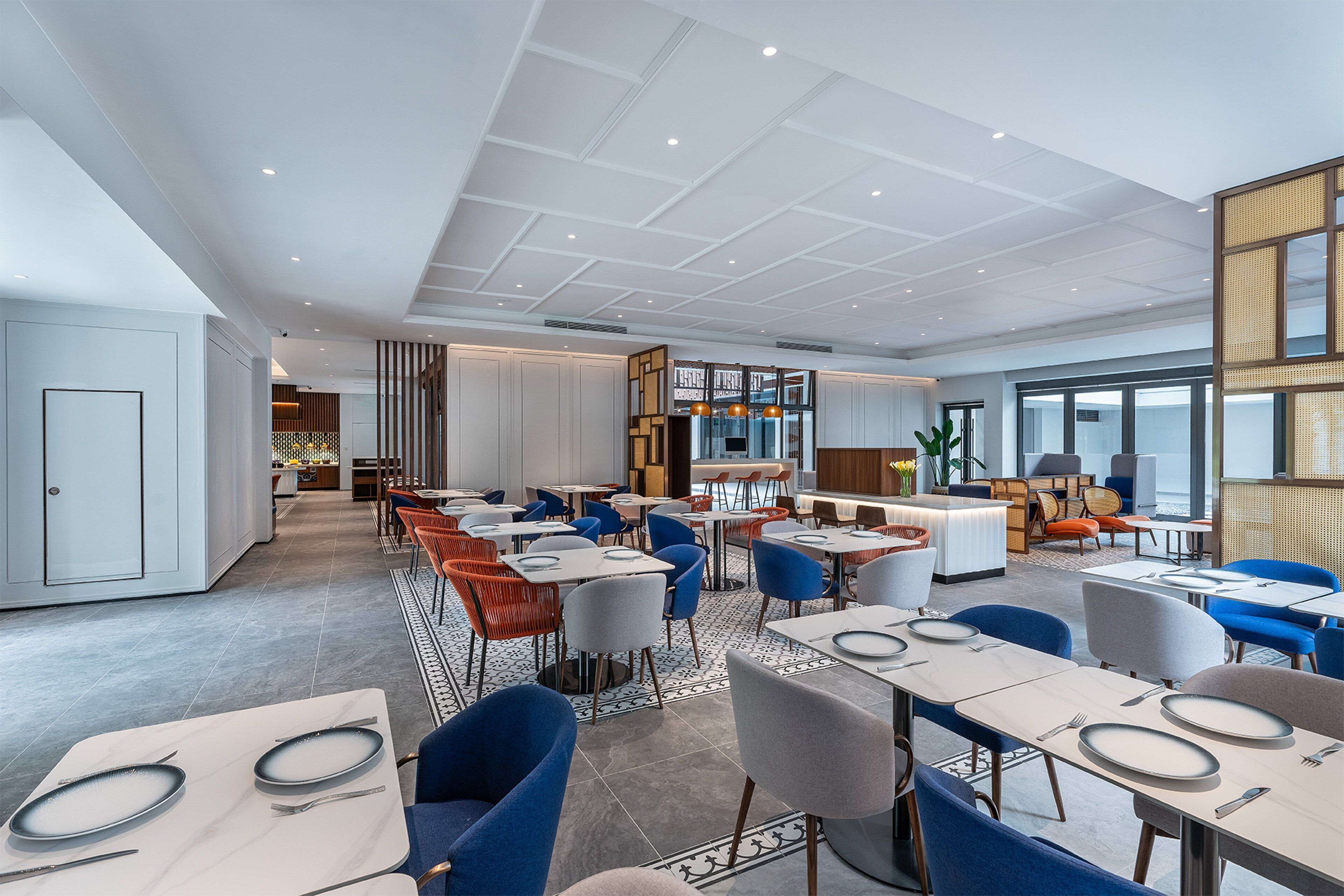A modern dining area with multiple tables and chairs, a kitchen in the background, and a large window providing natural light.