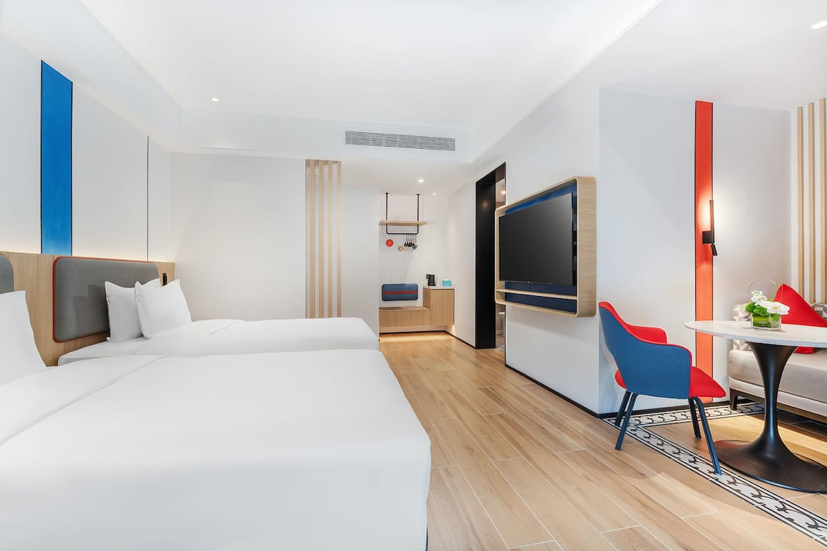 A modern hotel room with two beds, a TV, a small table with a chair, and a kitchenette.