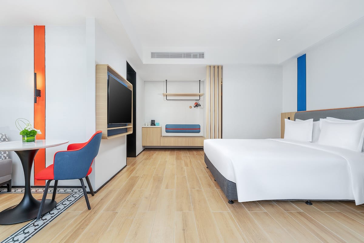 A modern hotel room with a bed, a desk with a chair, a TV, and a small table with a plant.