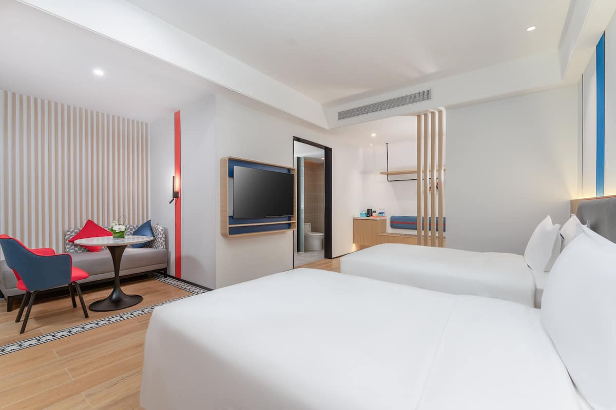 A hotel room with two beds, a TV mounted on the wall, a small table with a chair, and a sofa with pillows.