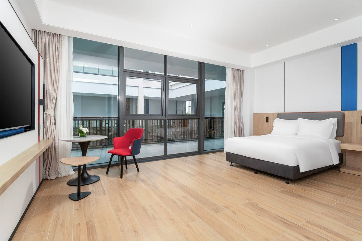A modern hotel room with a large bed, a TV, a small table with a chair, and a balcony with a view of the building.