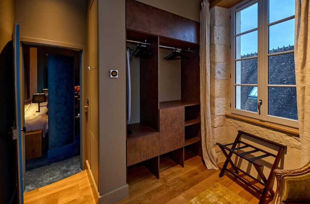 A room with a wooden floor, a bed with a blue headboard, a wardrobe, a window with curtains, and a small table.