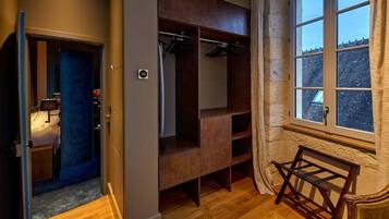 A room with a wooden floor, a bed with a blue headboard, a wardrobe, a window with curtains, and a small table.