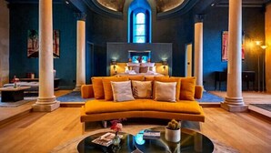 A luxurious living room with a large orange sofa, a coffee table, and a fireplace. The room has high ceilings, large windows, and classical columns. - La Chapelle, the only 5-star Suite in Nevers (Nevers)