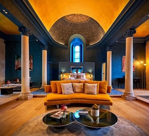 Ein luxuriöses Wohnzimmer mit einem großen orangefarbenen Sofa, einem Couchtisch und einem Kamin. Das Zimmer zeichnet sich durch hohe Decken, große Fenster und klassische Säulen aus.