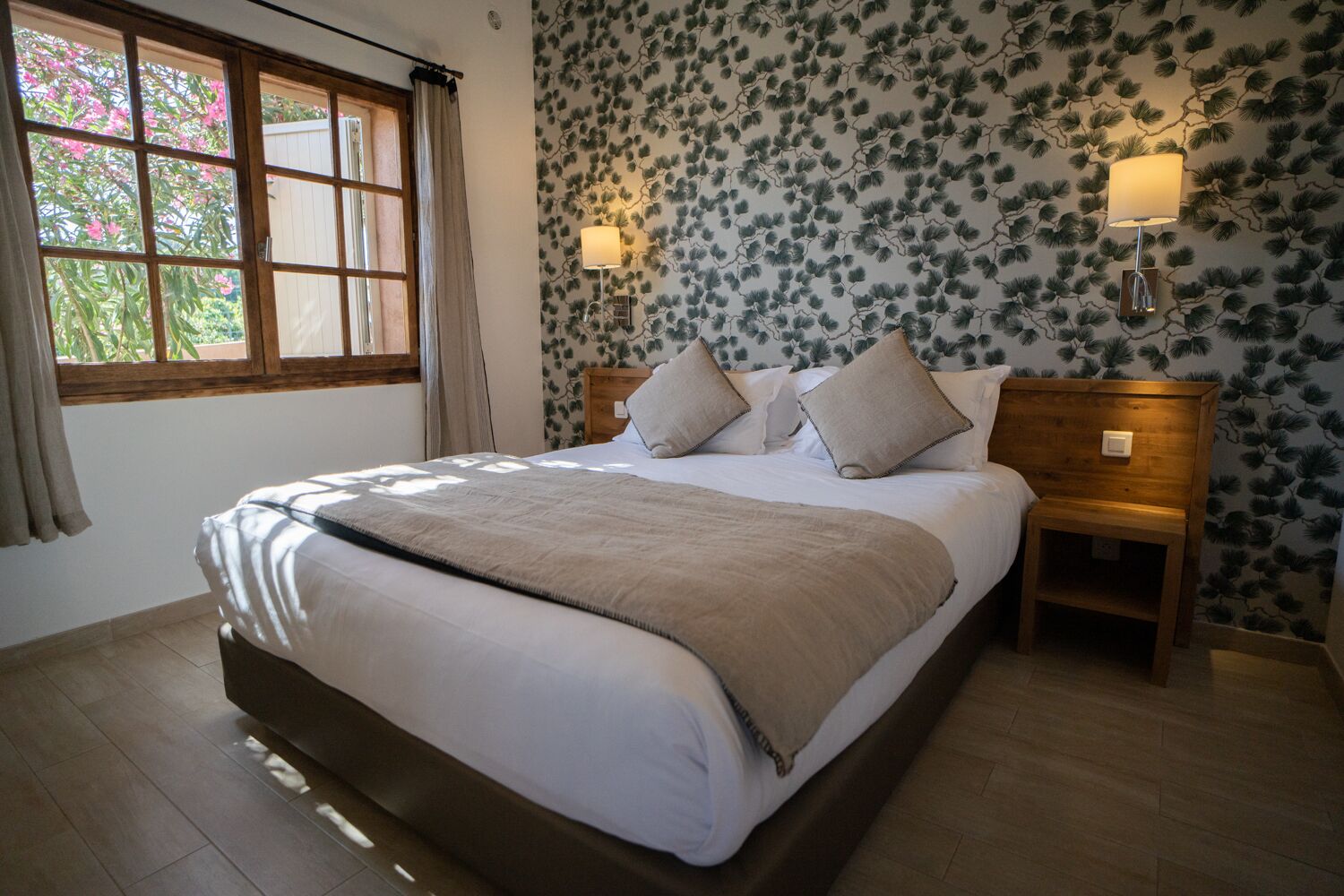 A bedroom with a large bed, two bedside tables, a window with curtains, and a wall with a leafy pattern.